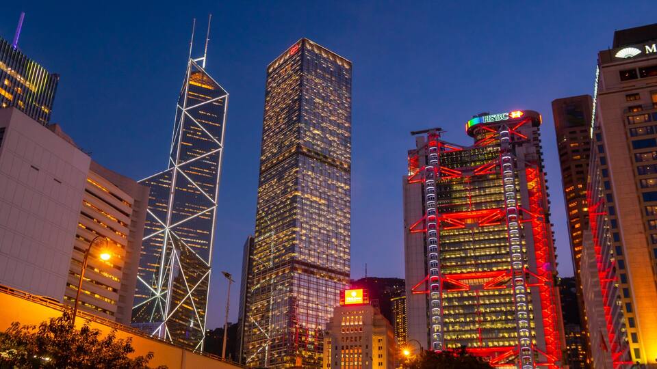 Une vue sur une ville au crépuscule, avec des gratte-ciel illuminés, dont un bâtiment remarquable à la structure squelettique blanche et un autre éclairé en rouge.