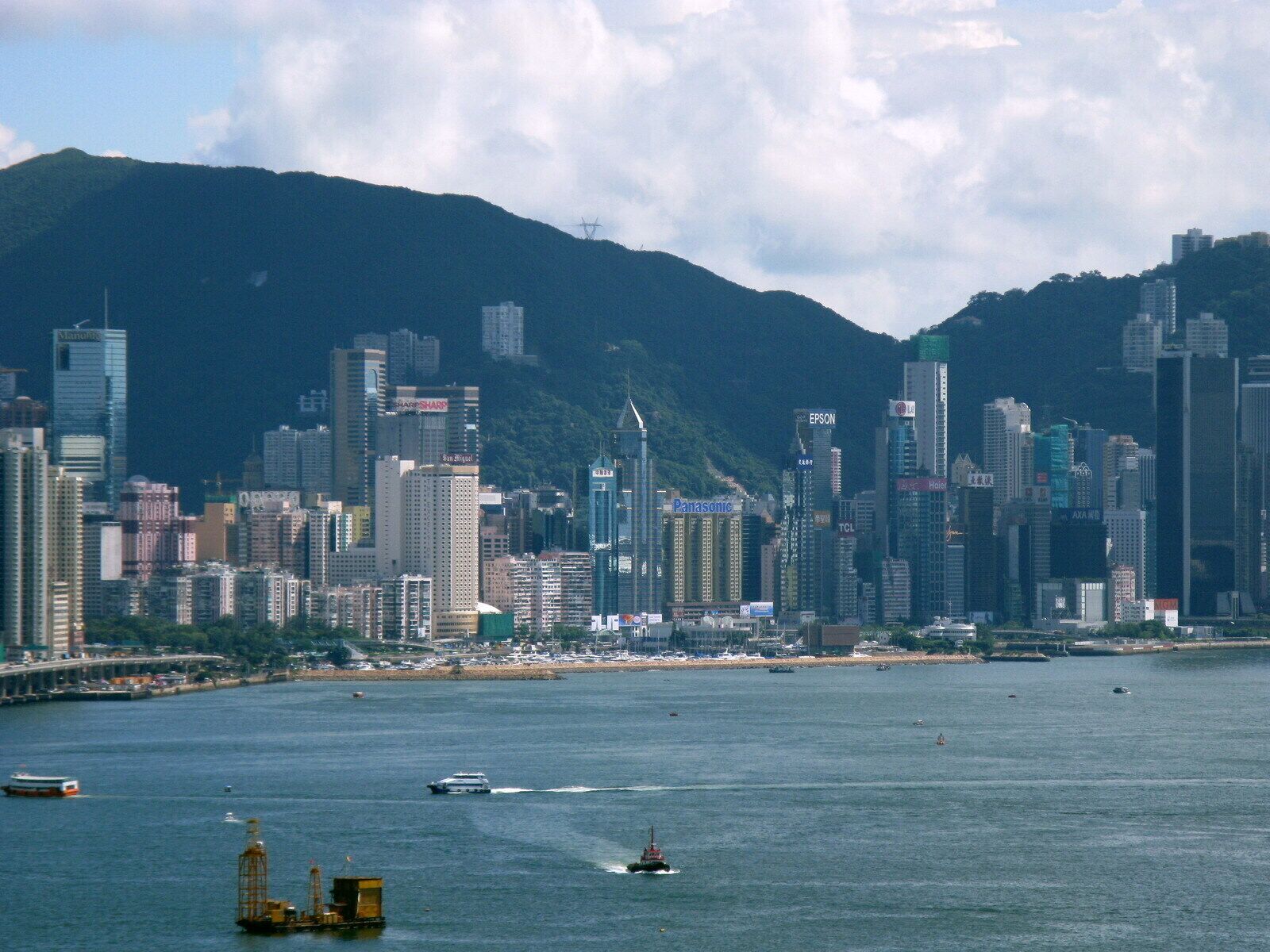 A city skyline with high-rise buildings, a body of water with boats, and a mountainous backdrop.