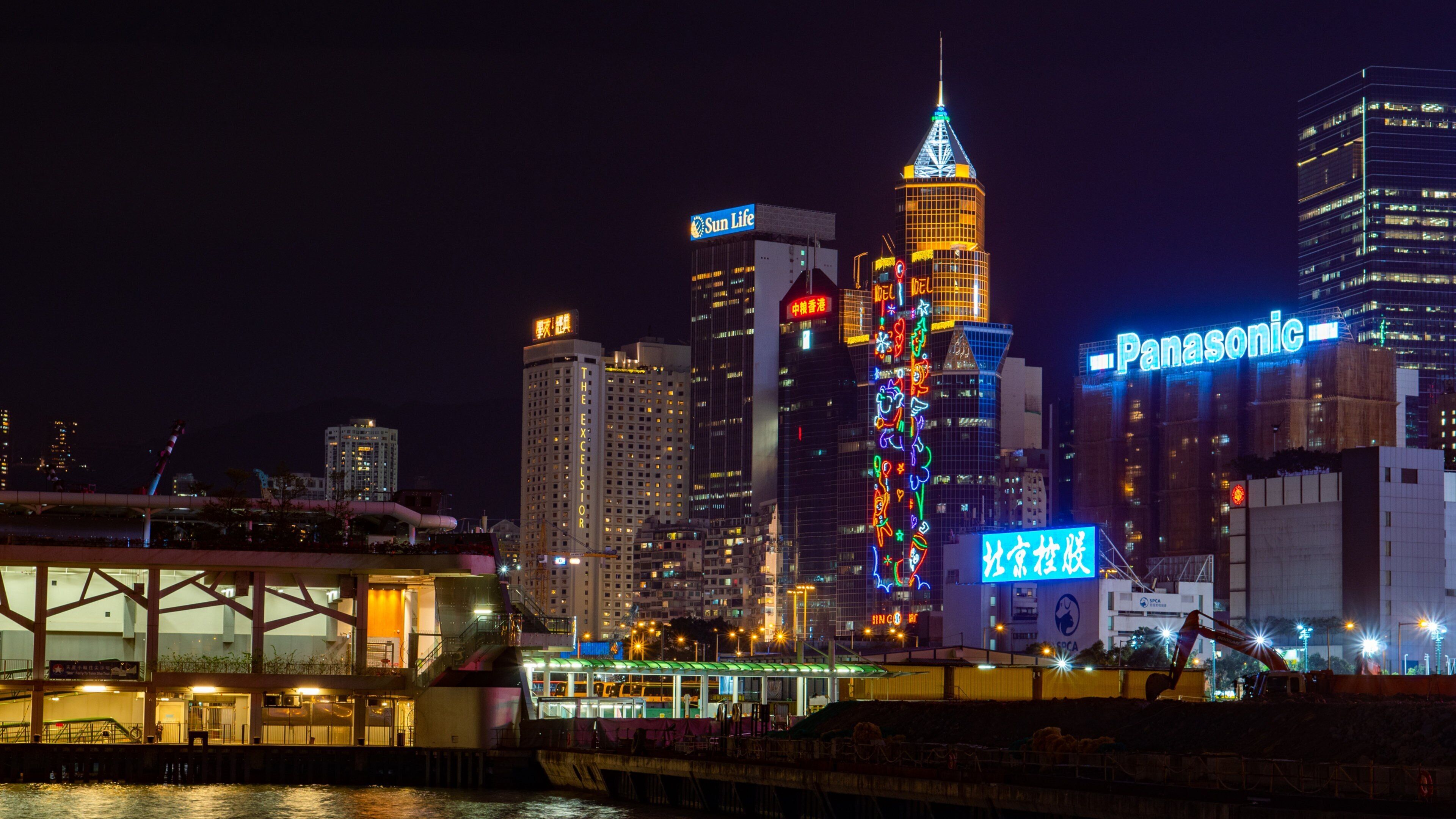 A city skyline at night with illuminated buildings, including a prominent tower with a pointed top and a building with a Panasonic sign.