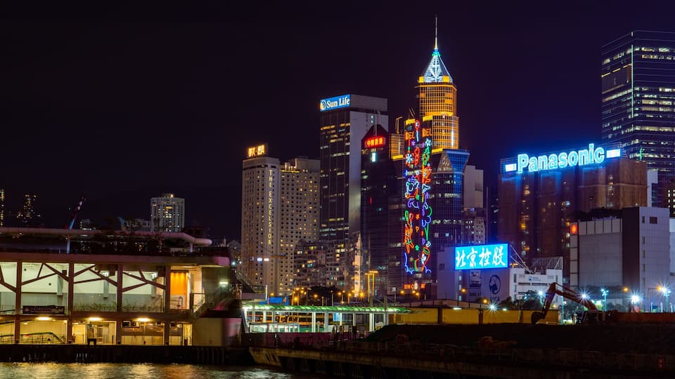 A city skyline at night with illuminated buildings, including a prominent tower with a pointed top and a building with a Panasonic sign.