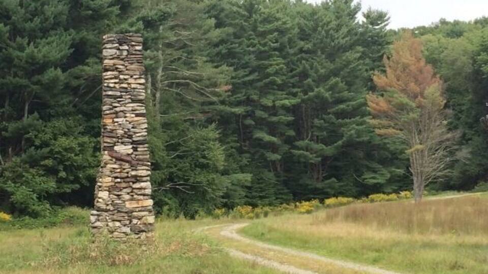 A stone structure in a grassy field with a dirt path leading to a forest.