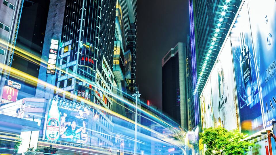 A nighttime cityscape with tall buildings, illuminated billboards, and light trails from moving vehicles.