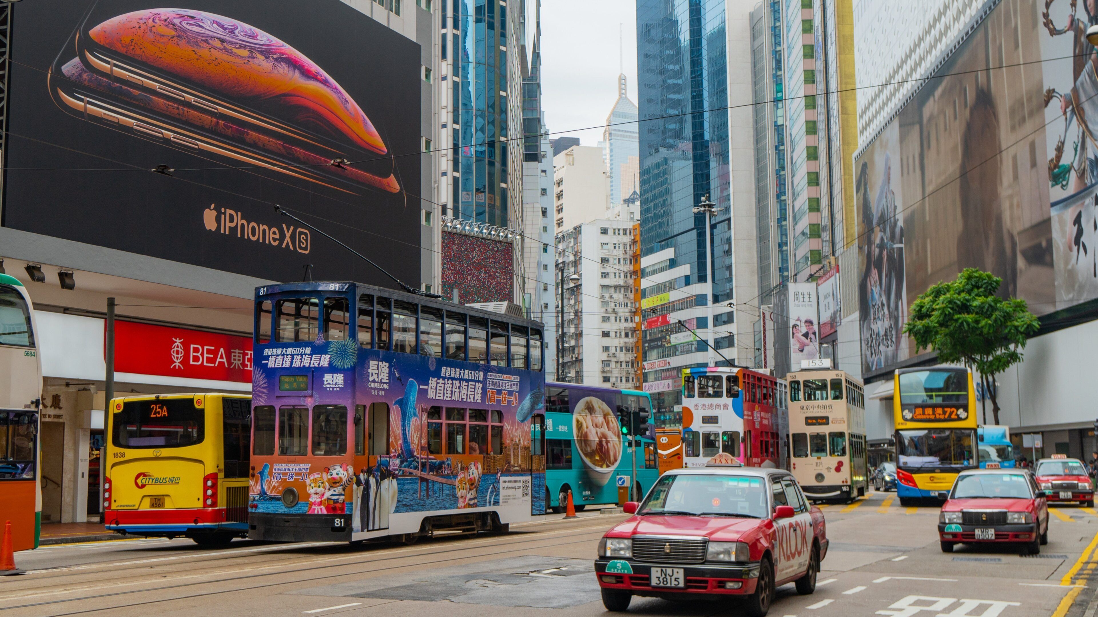 A busy urban street with double-decker buses, taxis, and a large Apple iPhone X advertisement on a building.