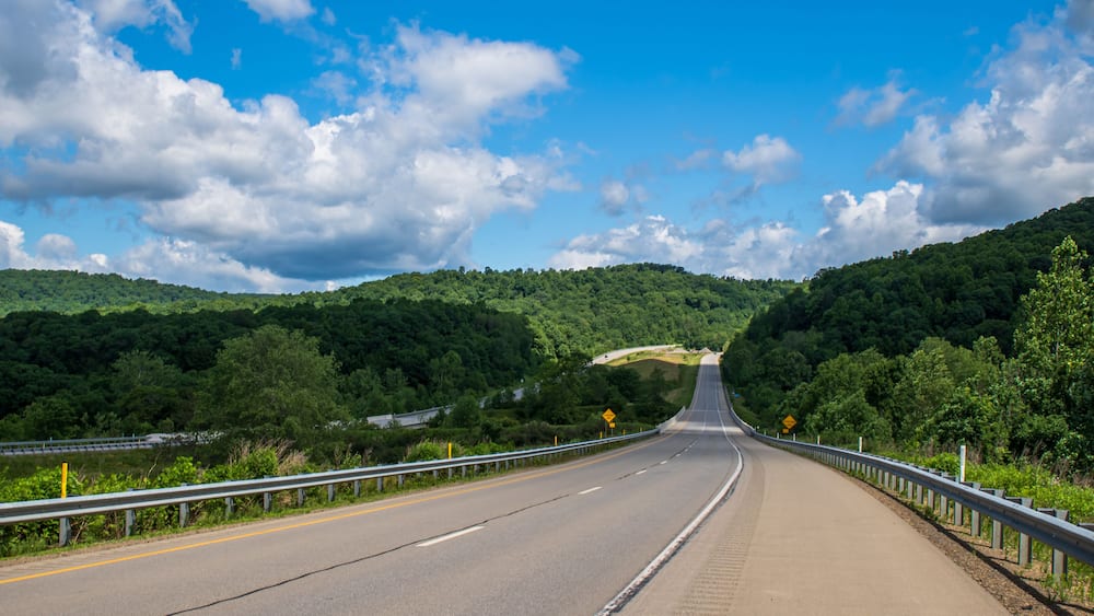 Eine kurvenreiche Autobahn führt durch ein bewaldetes Tal, gesäumt von einer Schutzplanke, unter blauem Himmel.