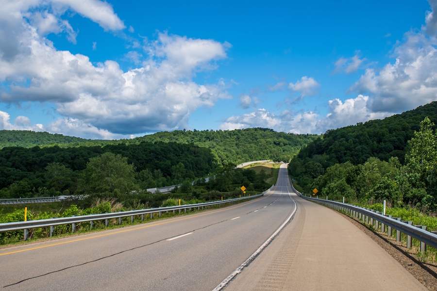A highway curving through a forested valley with a guardrail and a clear blue sky.