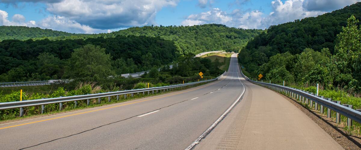 A highway curving through a forested valley with a guardrail and a clear blue sky.