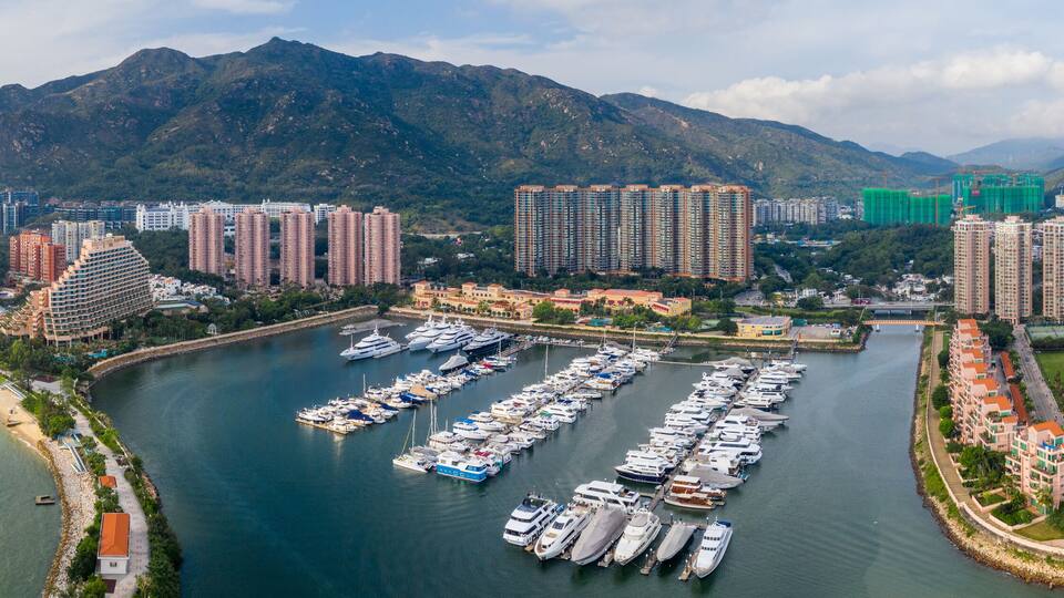 A marina with numerous boats docked, surrounded by high-rise residential buildings and a mountainous backdrop.
