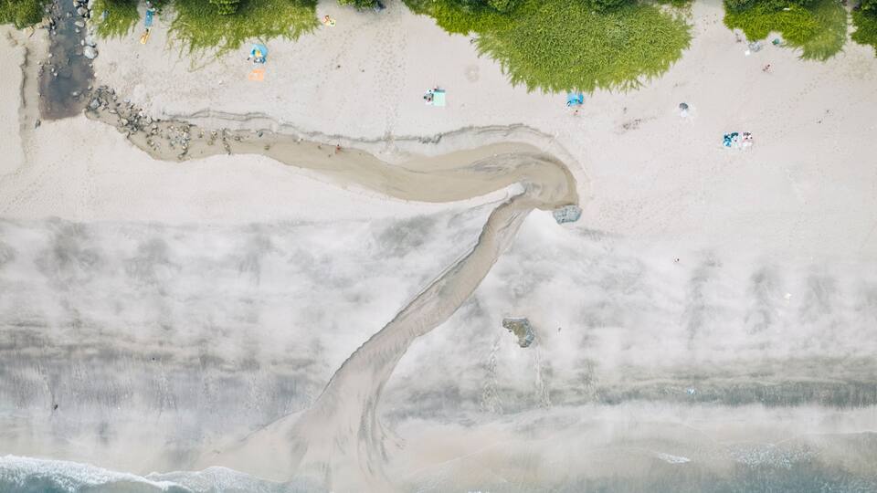An aerial view of a beach with a long sandbar extending into the ocean, surrounded by dense green forest.