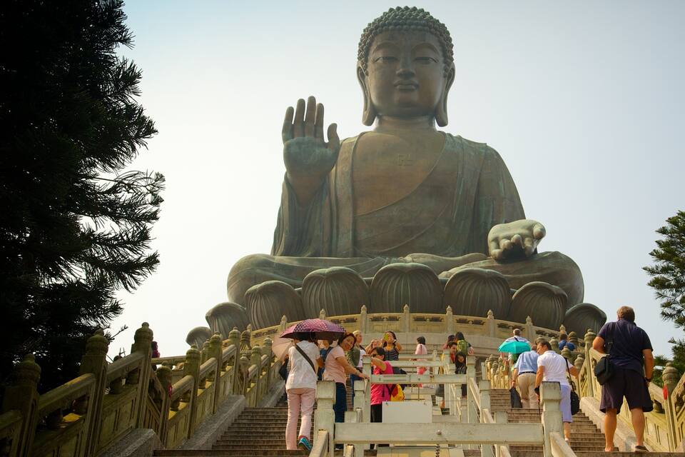 A large Buddha statue with a raised hand, surrounded by stairs and railings, with people ascending and descending.