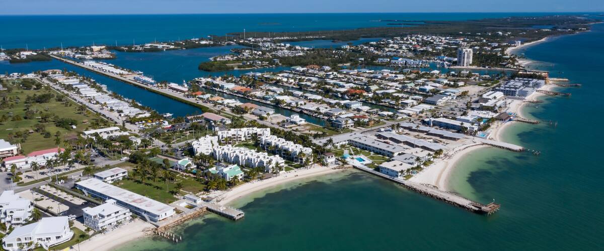 An aerial view of a coastal town with a marina, beachfront properties, and a clear blue sea.