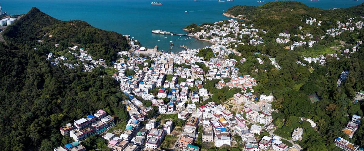An aerial view of a coastal town with dense housing, a harbor with boats, and a mountainous backdrop.