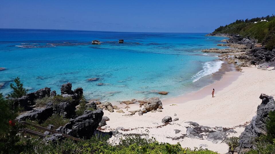 A beach with clear turquoise waters, rocky outcrops, and a person standing on the sand.