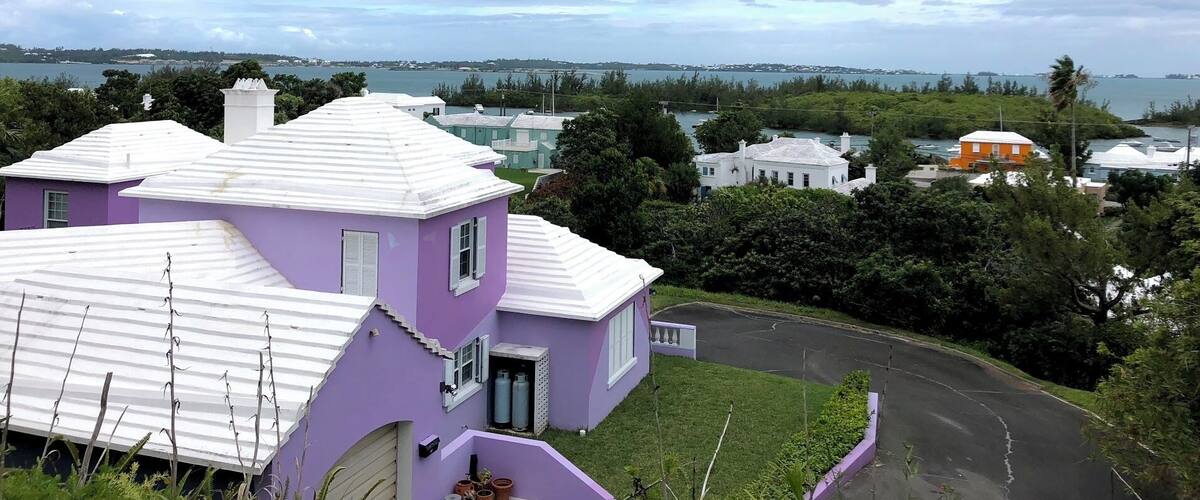 A purple house with a white roof, a parking lot, and a view of the sea and distant buildings.