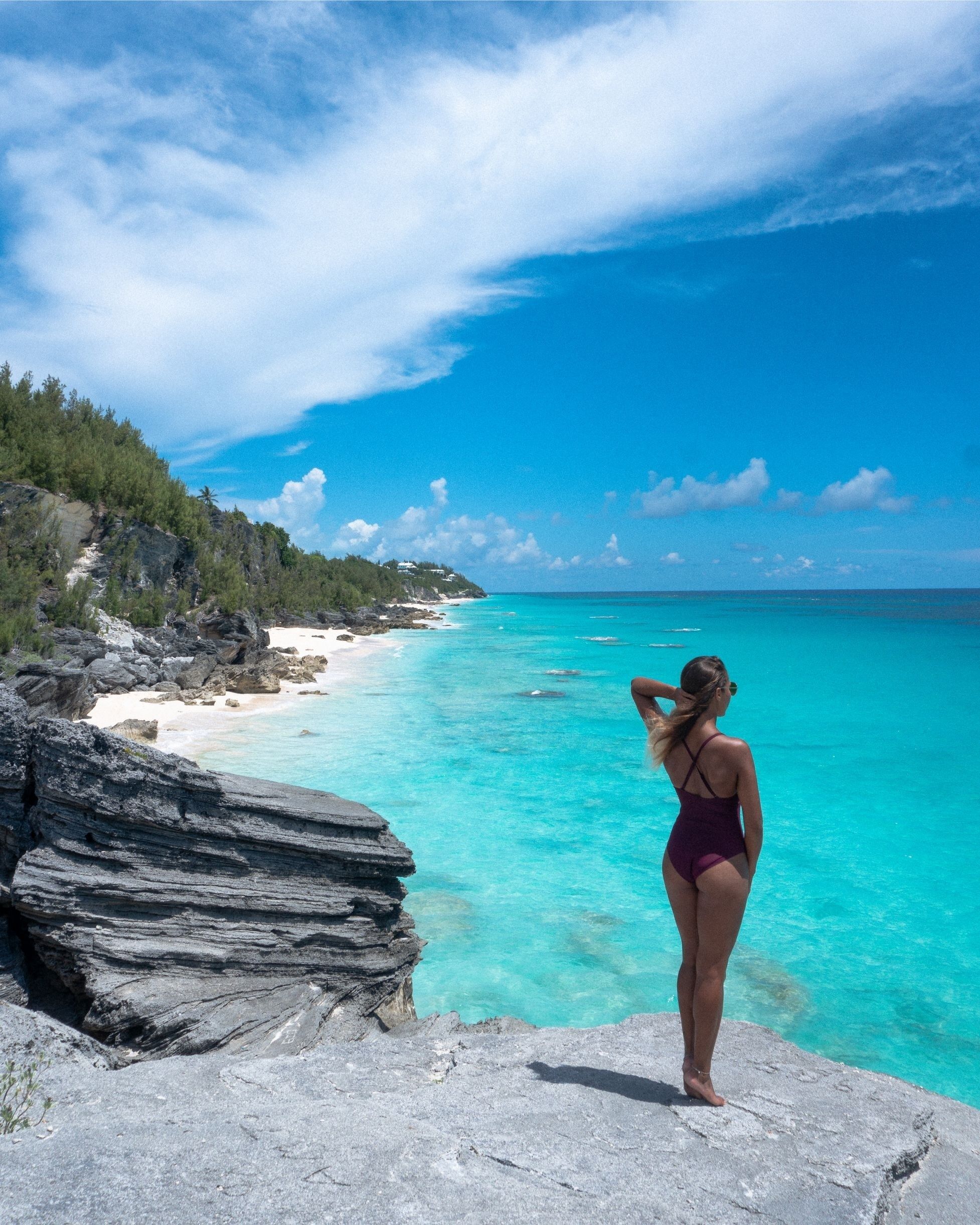 A woman in a purple swimsuit standing on a rocky outcrop overlooking turquoise waters and a forested coastline.