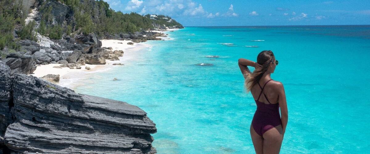 A woman in a purple swimsuit standing on a rocky outcrop overlooking turquoise waters and a forested coastline.