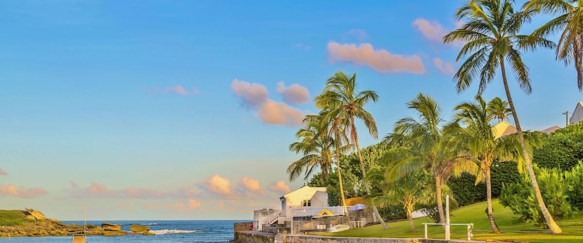 A calm waterfront with boats, palm trees, and a white building with a yellow roof.