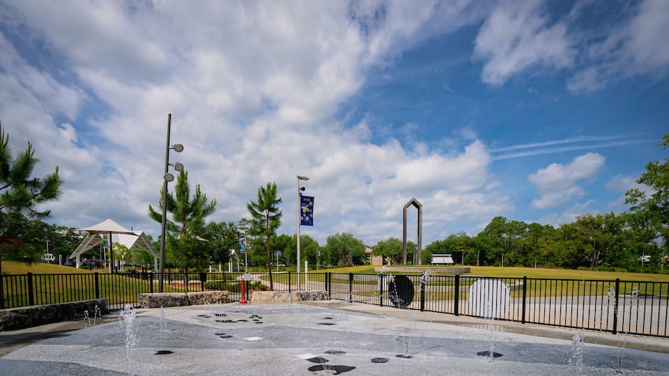 A park with a fountain, a flag, and a clear sky.