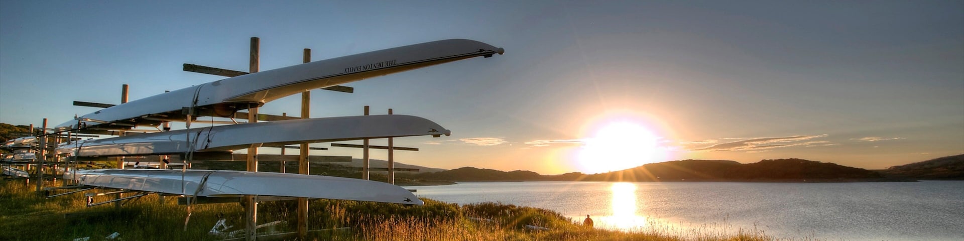 A sunset over a lake with several sailplanes parked on a grassy field.