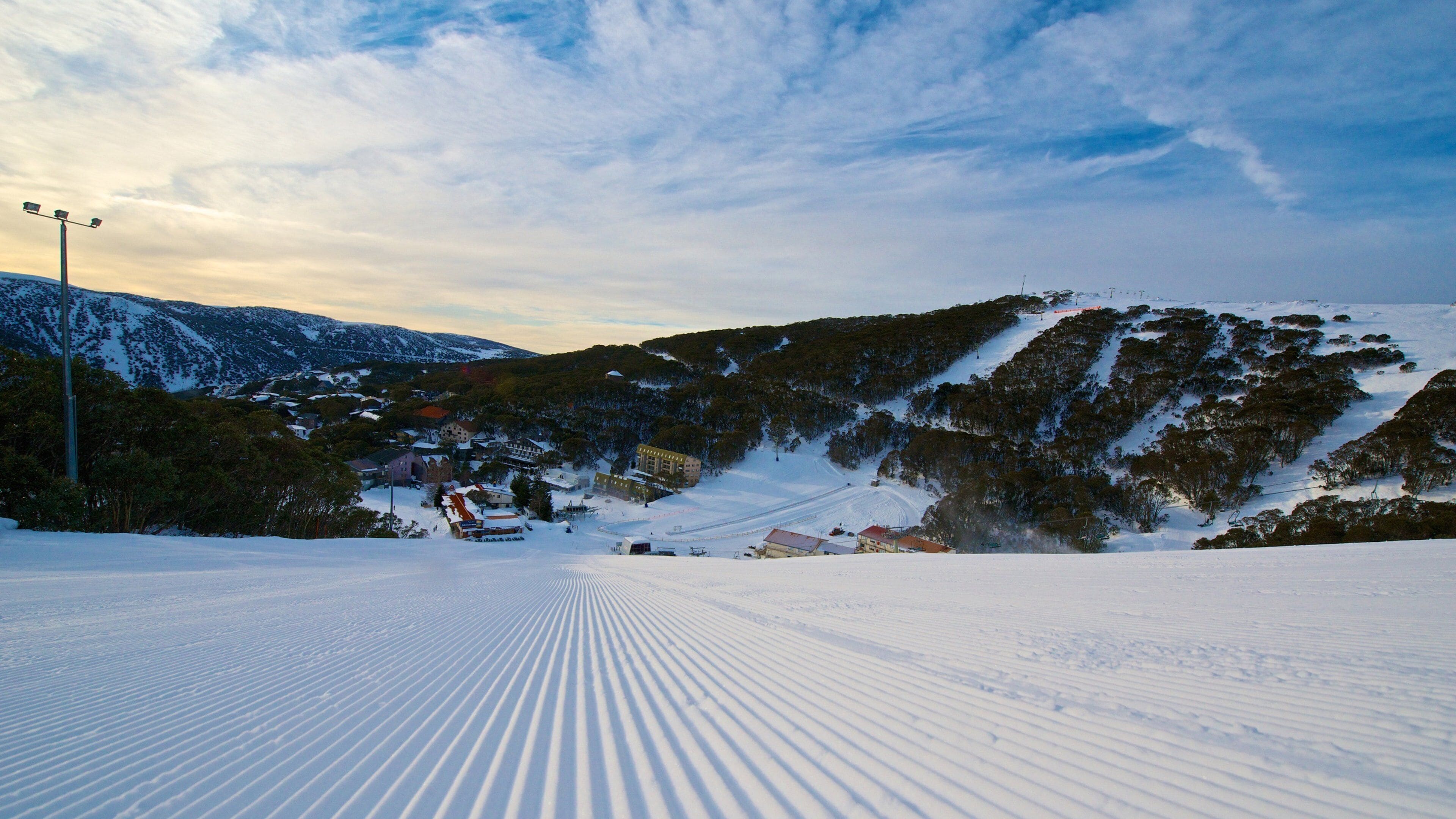 A snow-covered ski slope with a ski lift, buildings, and a forested mountain.