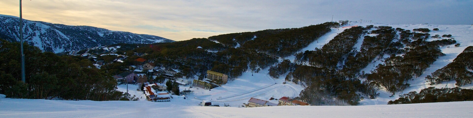 A snow-covered ski slope with a ski lift, buildings, and a forested mountain.