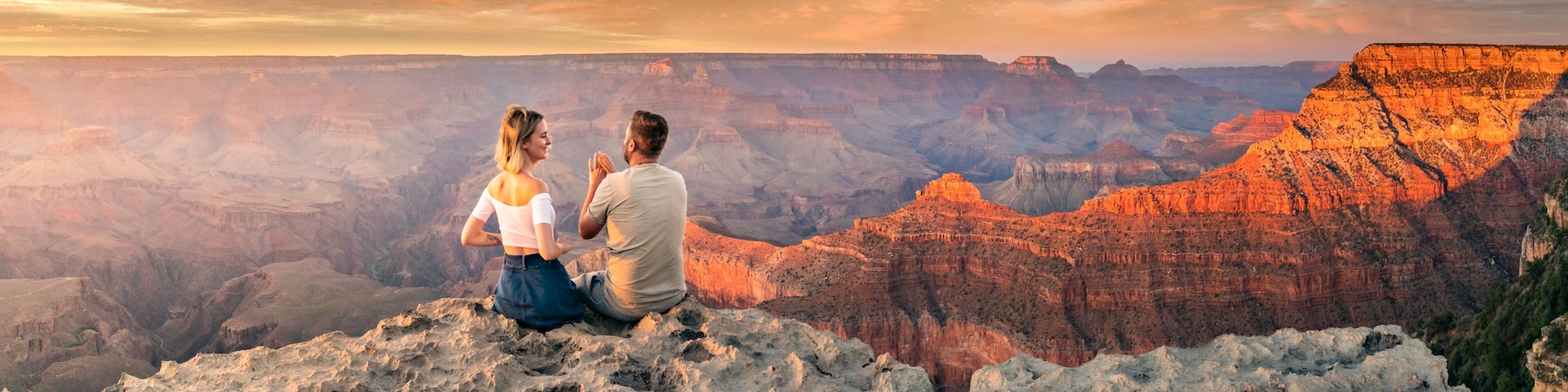 Una pareja sentada sobre un afloramiento rocoso, con vistas a un cañón con formaciones rocosas en capas.
