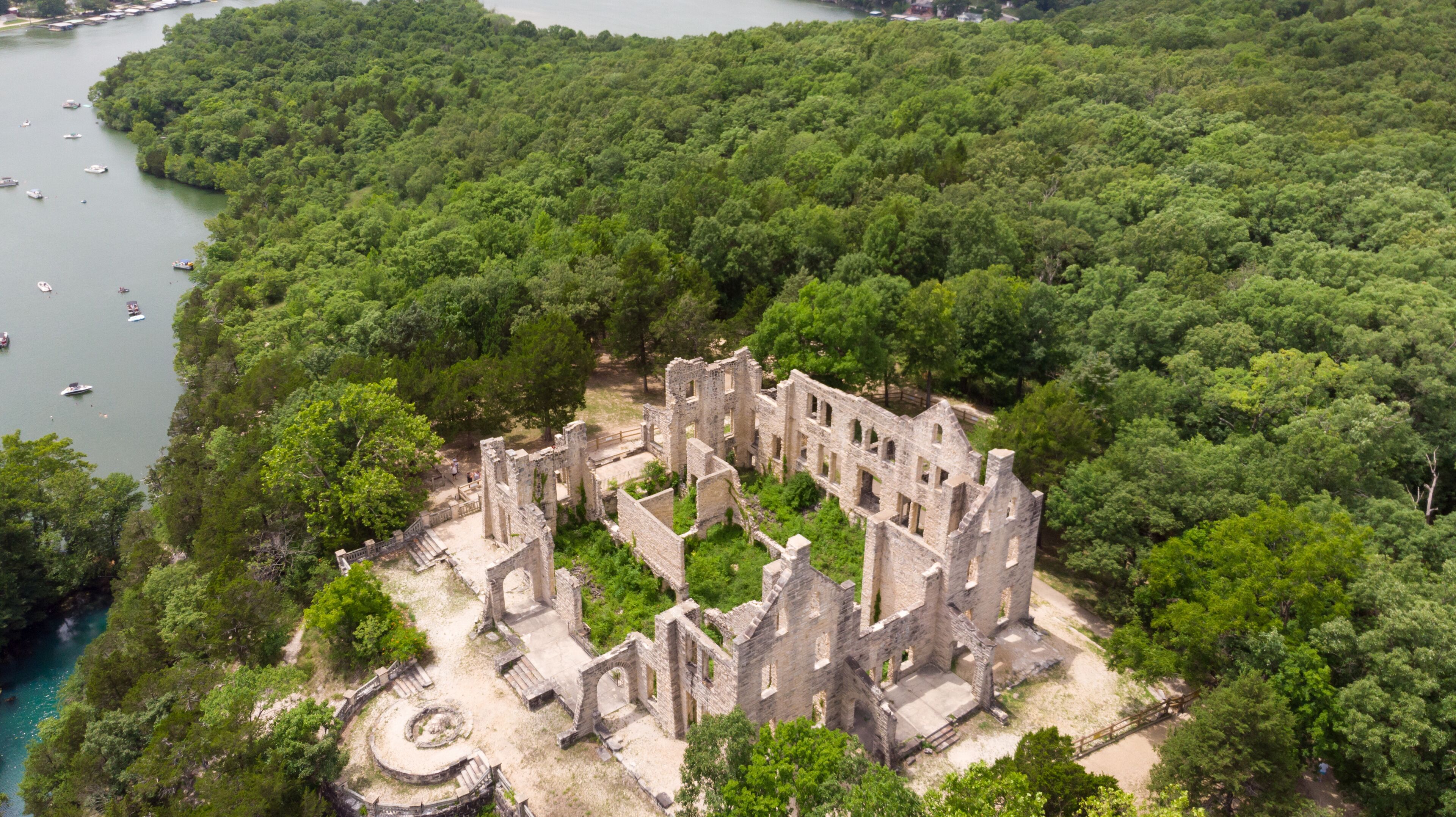 An aerial view of a historic stone ruin surrounded by dense forest, with a river in the background.