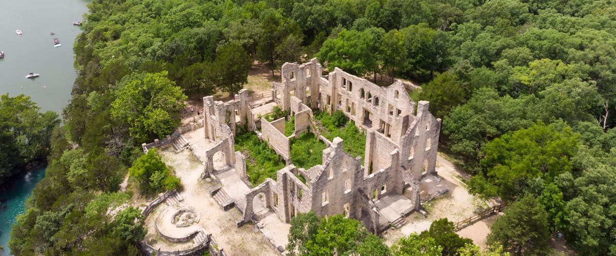 An aerial view of a historic stone ruin surrounded by dense forest, with a river in the background.