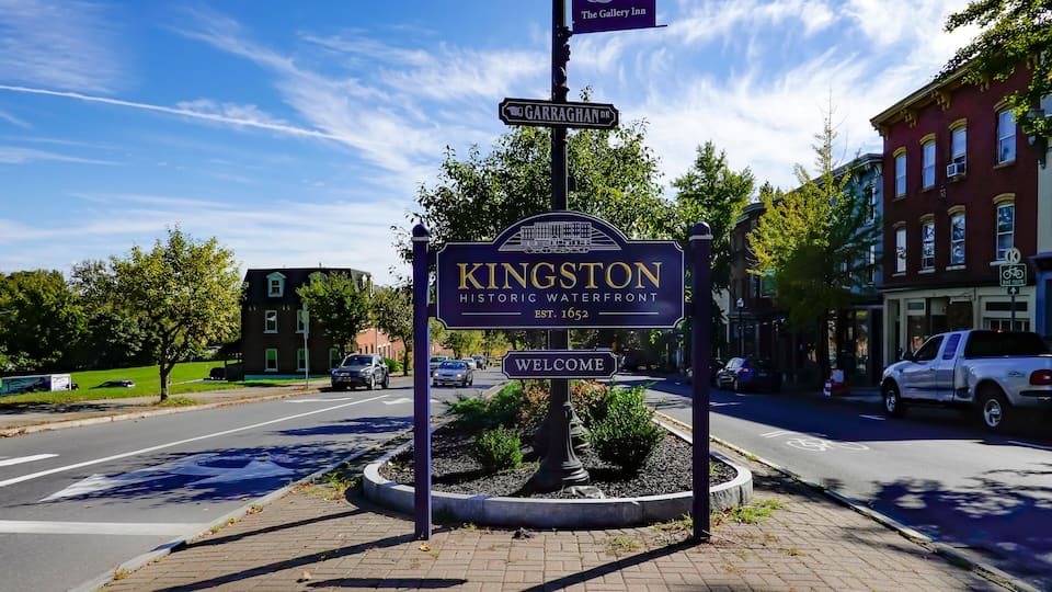 A street sign for Kingston Historic Waterfront with a welcome message, a Garraghan Dr. sign, and a The Gallery Inn sign.