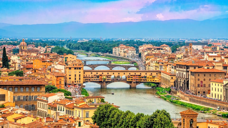 A river with bridges, historic buildings, and a mountainous backdrop.