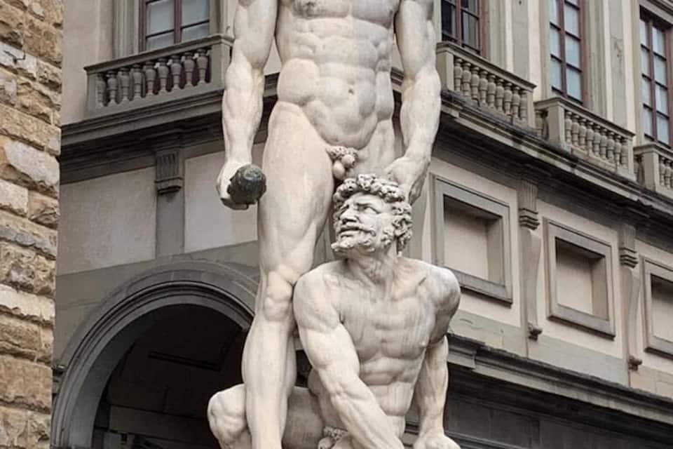 A classical statue of a standing man and a kneeling man with a lion, set against a building with arched windows and columns.