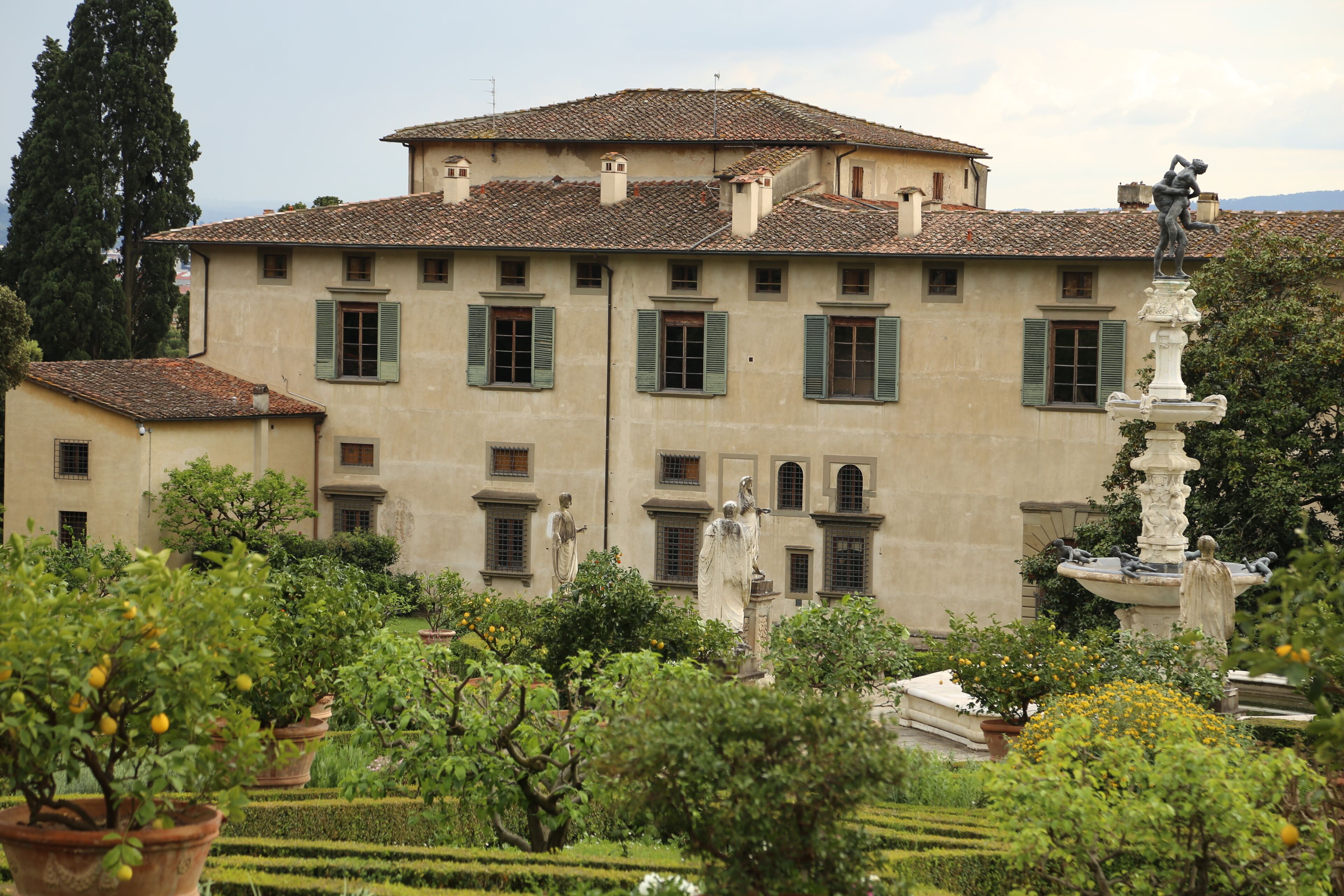 A historic building with green shutters, a fountain, and statues in a well-manicured garden.