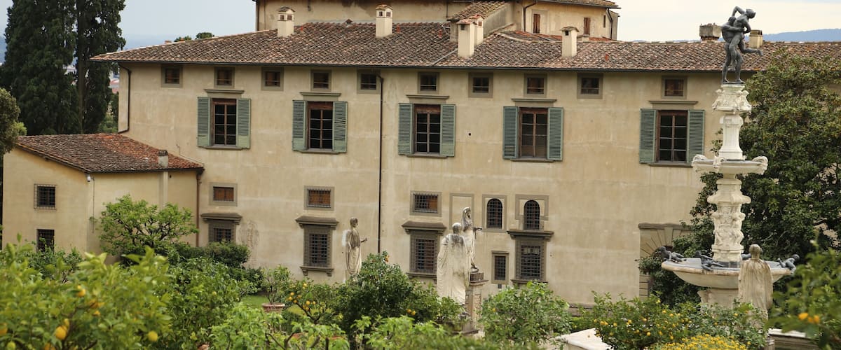 A historic building with green shutters, a fountain, and statues in a well-manicured garden.