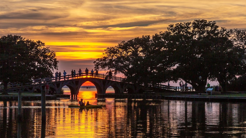 A sunset over a river with a bridge, trees, and people on the bridge.