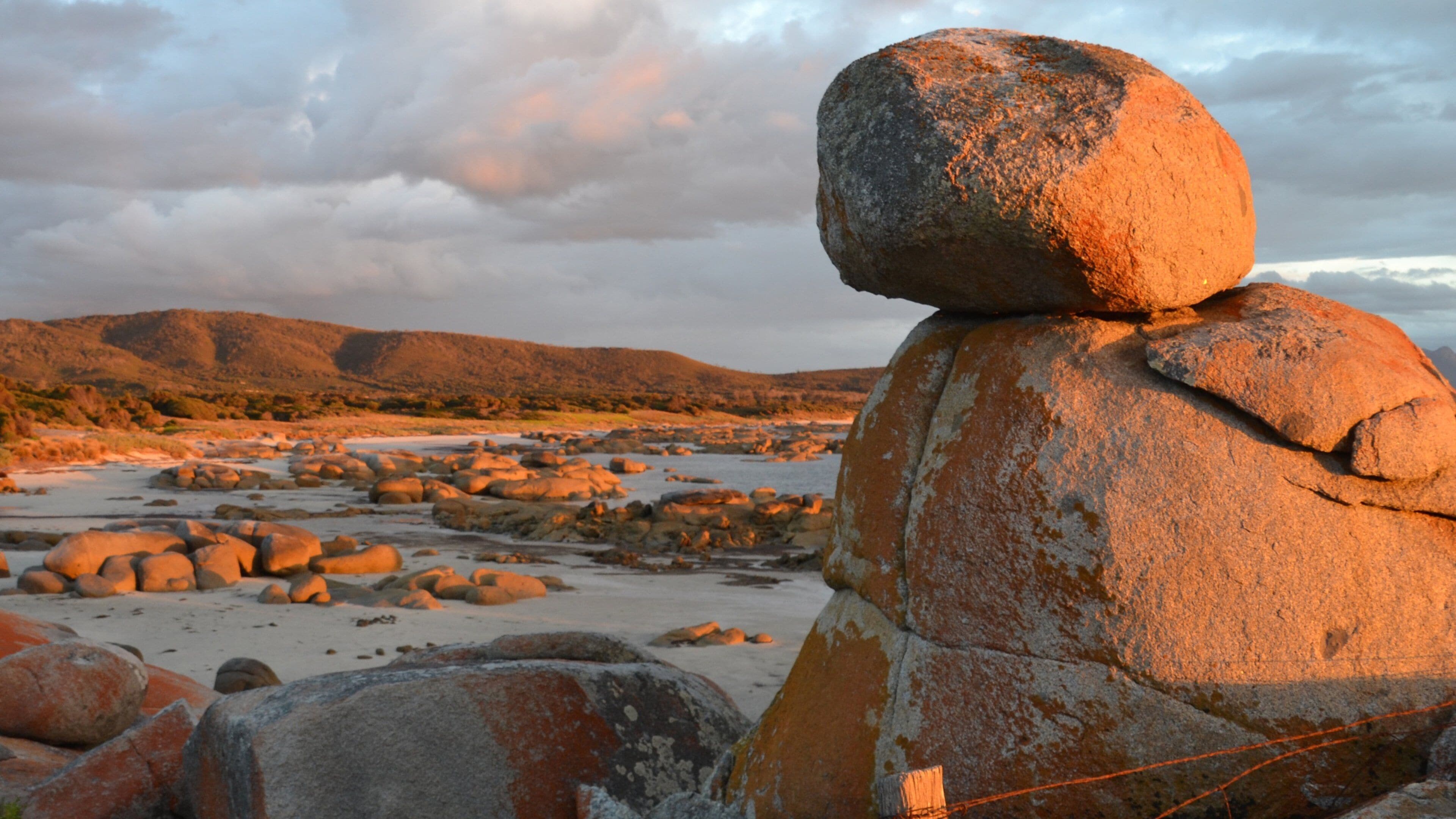 A large boulder balanced on another boulder in a rocky landscape with distant hills.