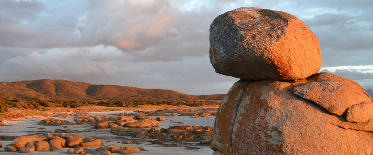 Una gran roca equilibrada sobre otra roca en un paisaje pedregoso con colinas distantes.