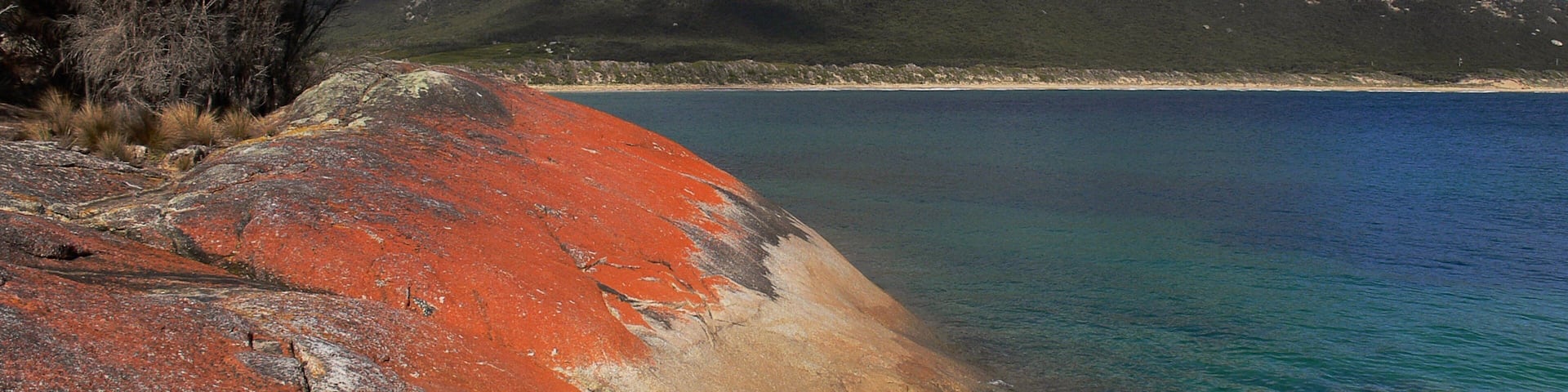 A coastal landscape with a rocky shoreline, a clear blue sea, and a mountain in the background.