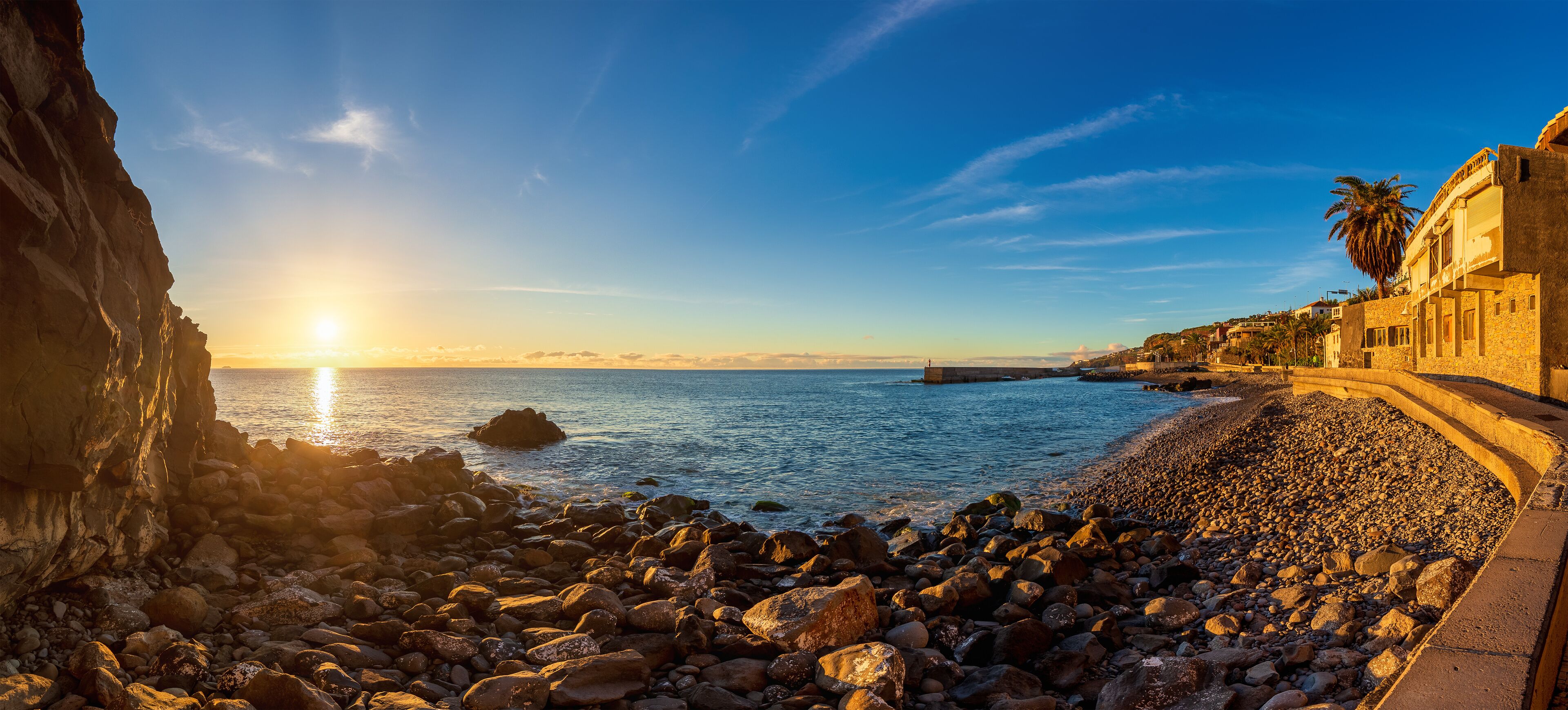 A rocky coastline with a sunset, a lighthouse, and buildings along the shore.