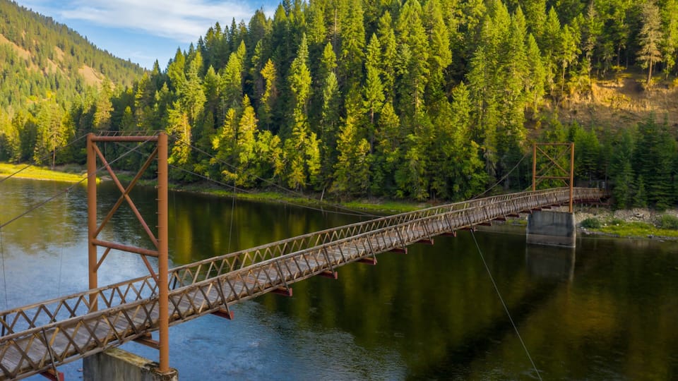 A suspension bridge over a calm river, surrounded by dense forest and hills.