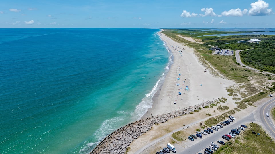A coastal road with a beach, parked cars, and a clear blue ocean.