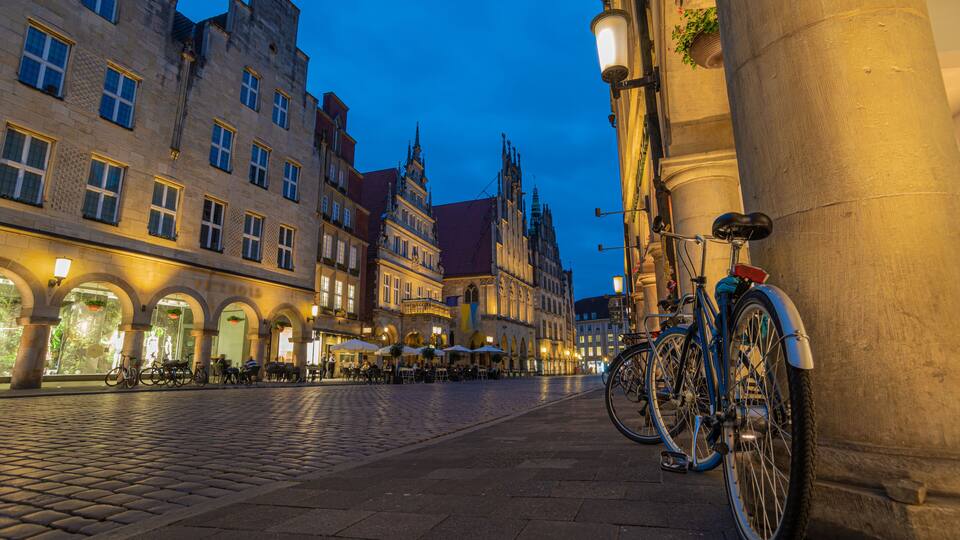 A cobblestone street lined with historic buildings, a bicycle parked against a column, and outdoor seating in the evening.