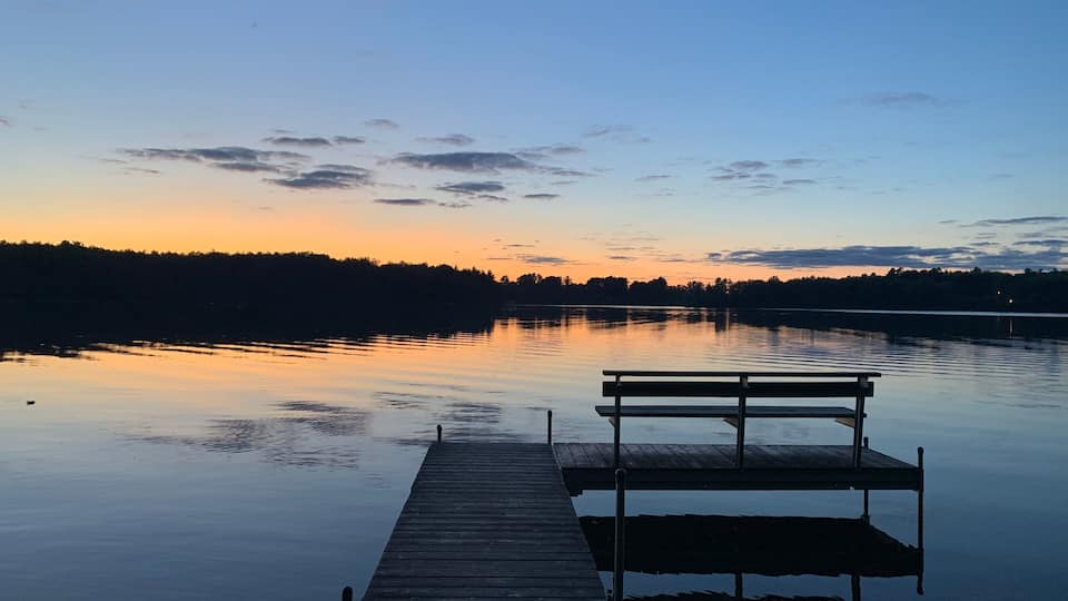 A wooden dock extending into calm water with a bench at the end, surrounded by a forested shoreline and a sunset sky.