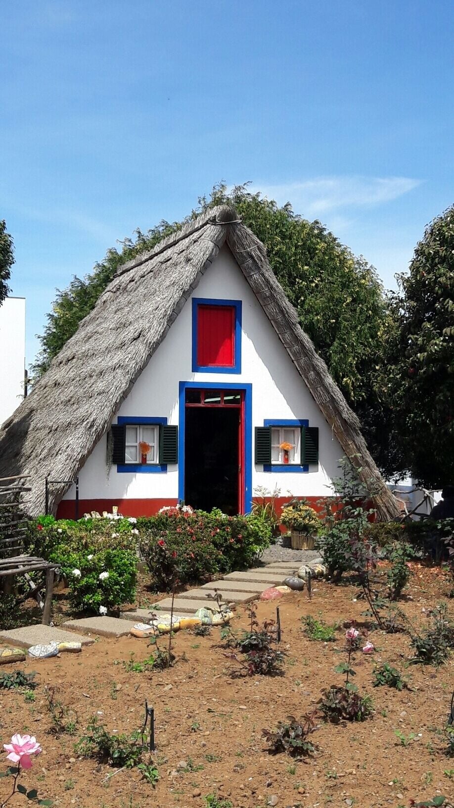 Una casa tradicional amb teulada de palla, porta vermella i persianes blaves, envoltada per un jardí amb plantes en flor i un camí de terra.