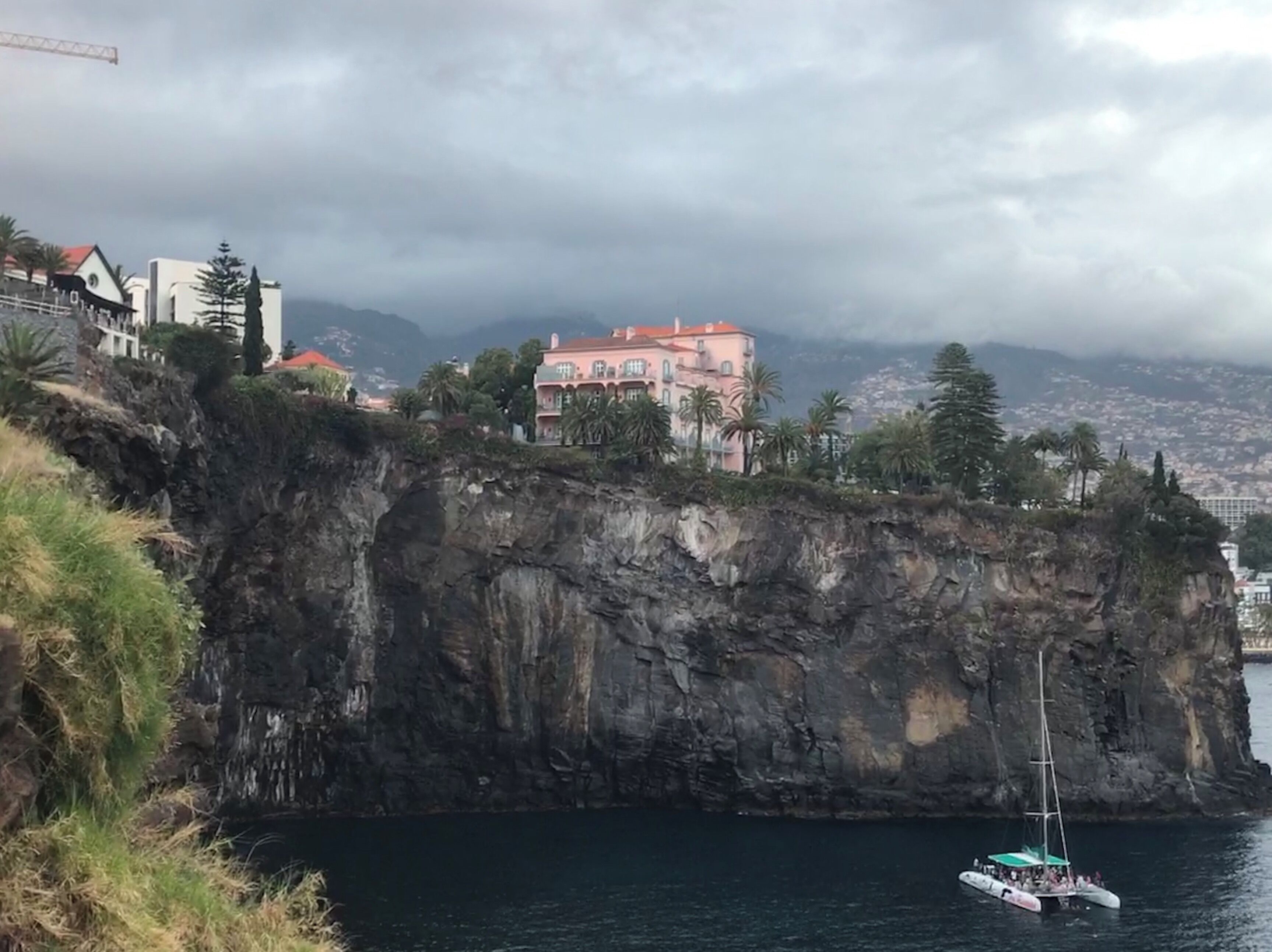 A coastal cliff with a pink house on top, a sailboat in the water, and a cityscape in the background.