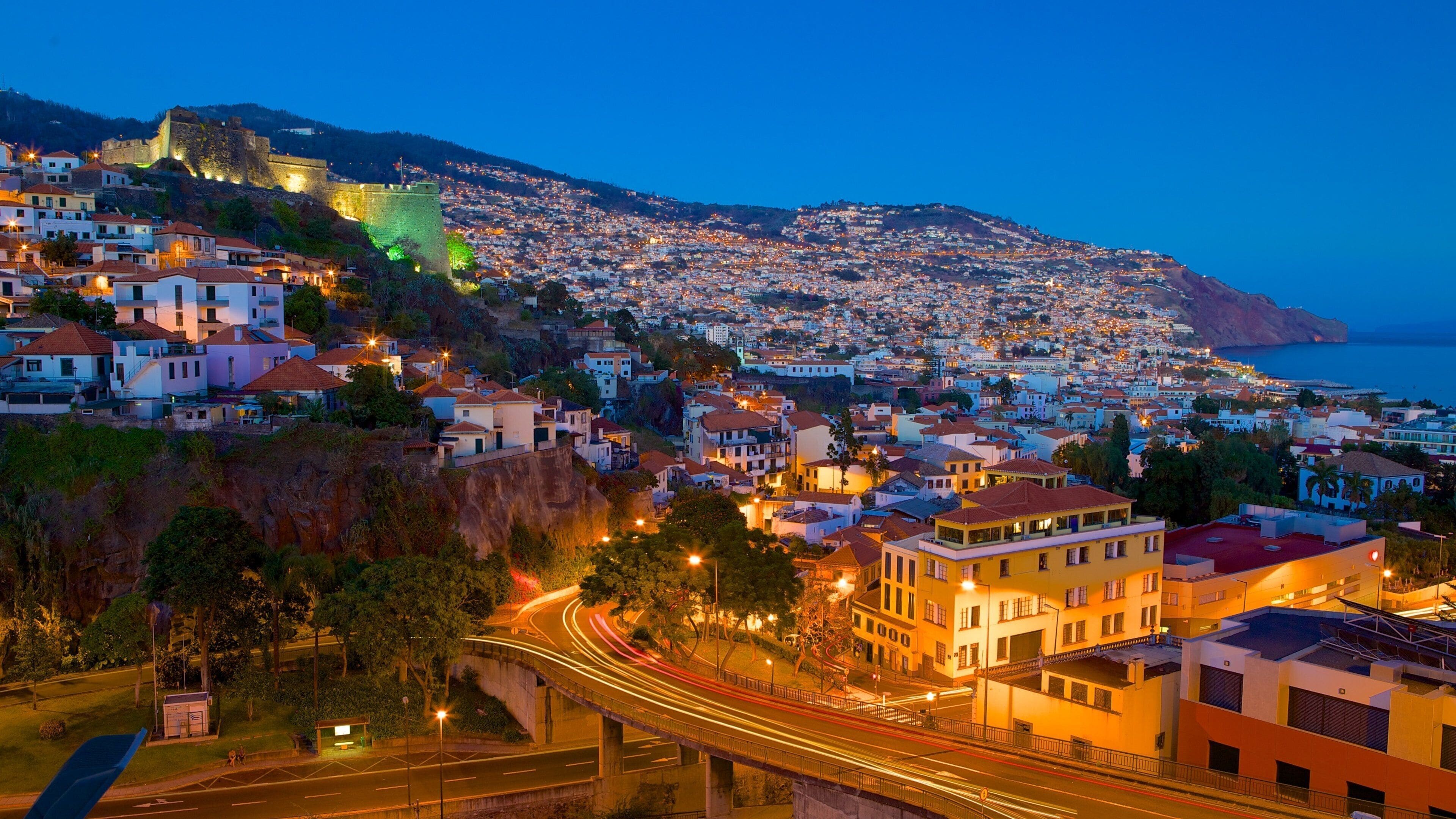 A coastal city at dusk with a prominent castle on a hill, illuminated streets, and a clear view of the sea.