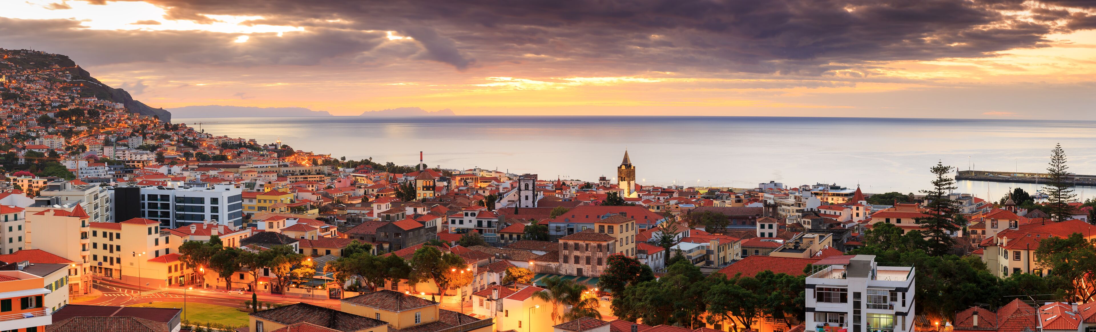 A coastal cityscape at sunset with a prominent church tower, a bridge, and a calm sea.