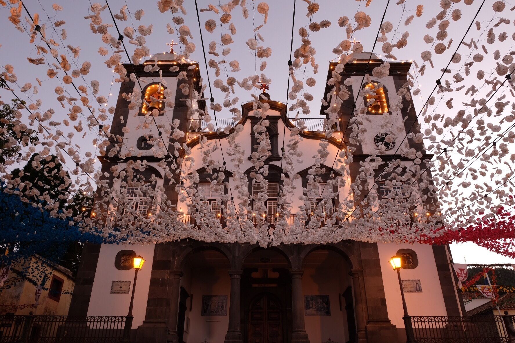 A church with two bell towers, decorated with white paper flowers and lights.