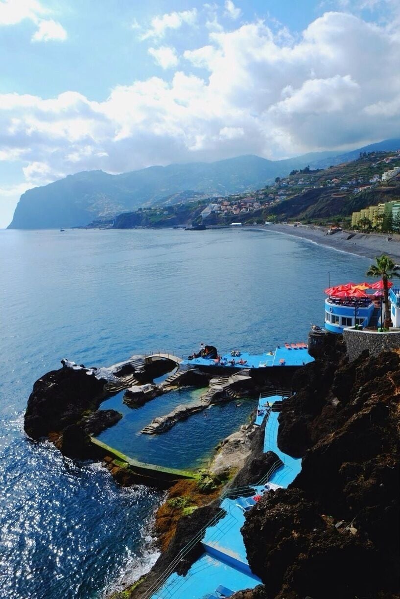 Un paisaje costero con costa rocosa, mar azul cristalino y una cadena montañosa distante.