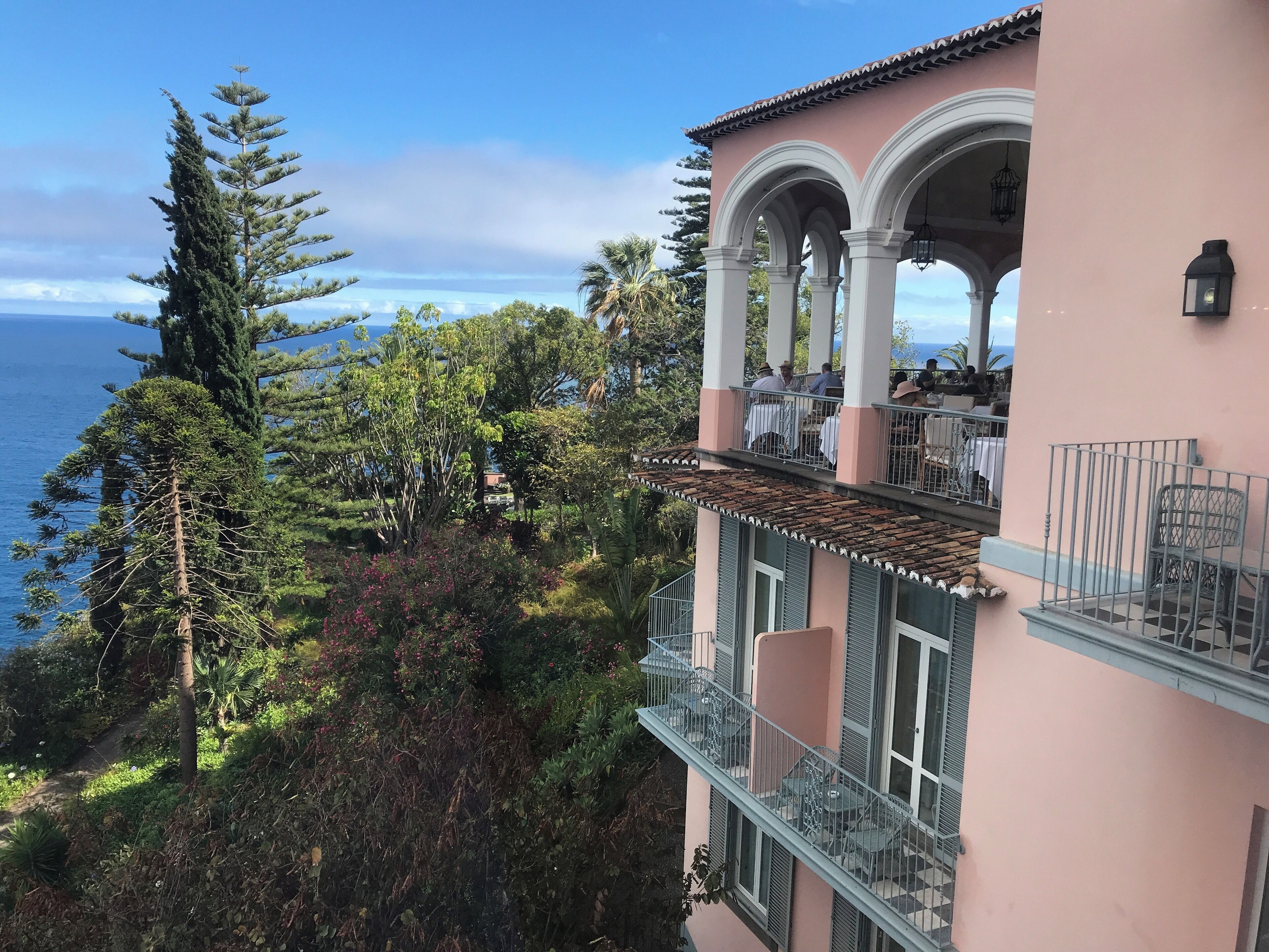 A coastal building with a balcony overlooking the sea, surrounded by trees and vegetation.