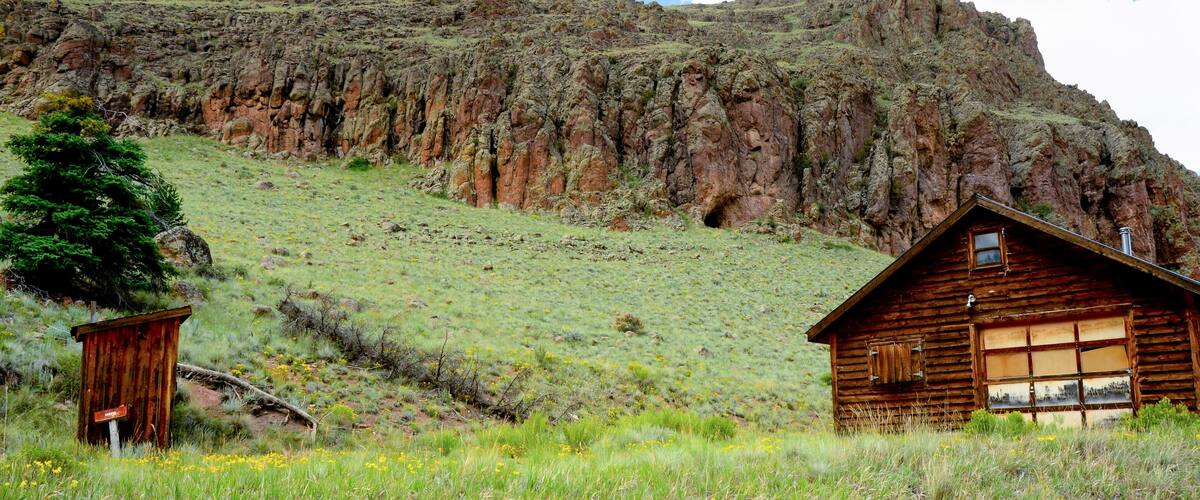 A wooden cabin on a grassy hillside with a rocky mountain in the background.