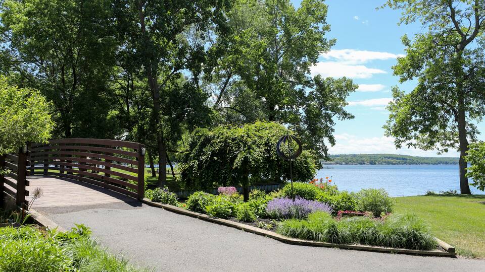 A wooden bridge over a paved path, surrounded by lush greenery and a lake.